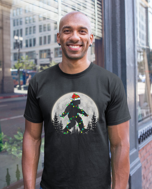 Black t-shirt featuring a Bigfoot silhouette wrapped in Christmas lights wearing a Santa hat under a full moon.
