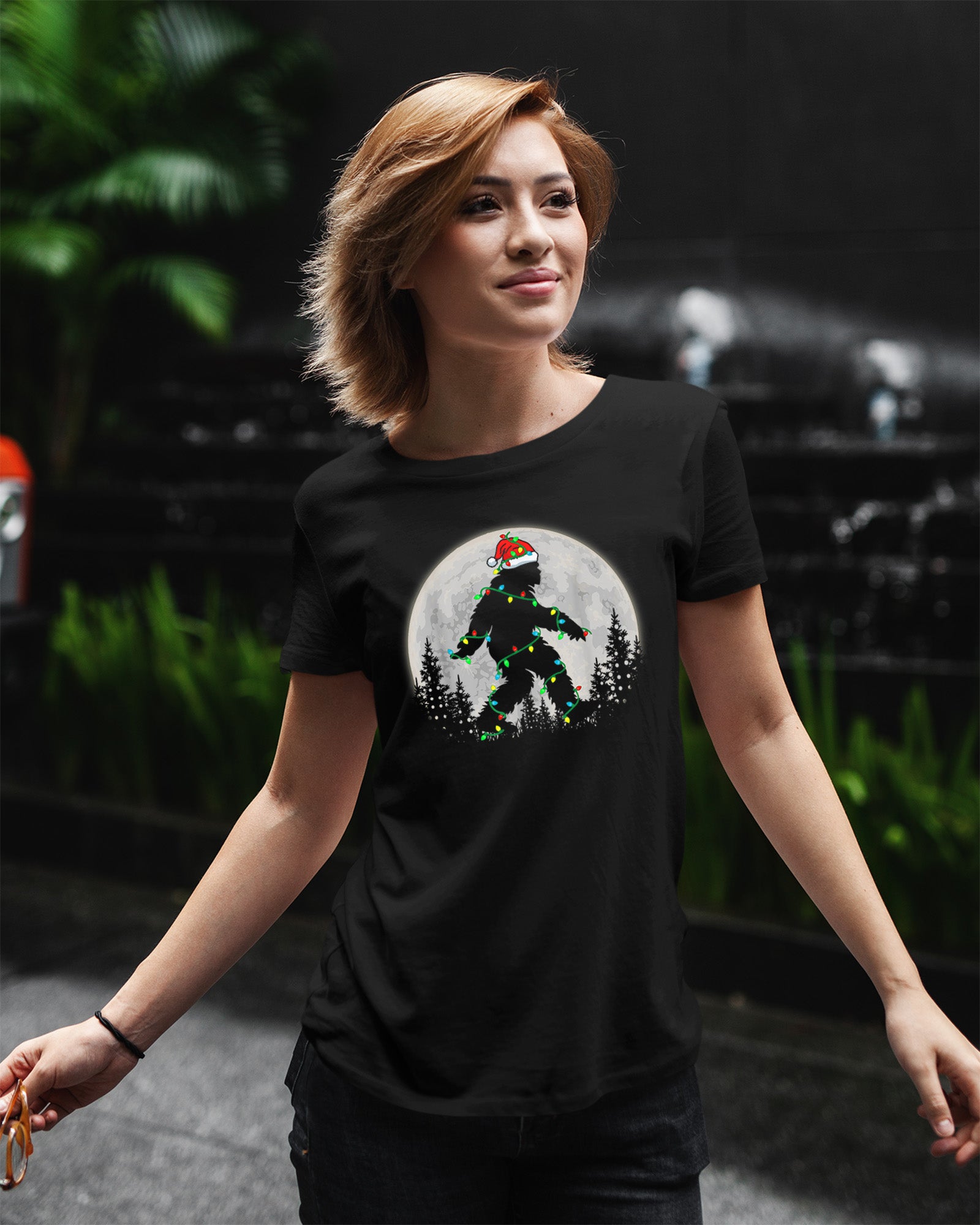 Black t-shirt featuring a Bigfoot silhouette wrapped in Christmas lights wearing a Santa hat under a full moon.